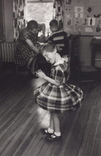 A young child smiles as they dance with eyes closed in the centre of this black and white photograph. The child is wearing a short sleeved plaid dress that billows up while they twirl. Behind them, an adult sits with a child at a table by a window.