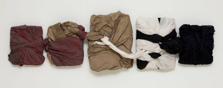 Five rectangles of roughly folded fabric are displayed in a horizontal line. From left to right the fabric overlaps from burgundy, beige, white and then black. The rectangles range in size, the ones on the outer edge the smallest, and the one in the centre, the largest. All of the rectangles are physically joined together by a tied strand of fabric.