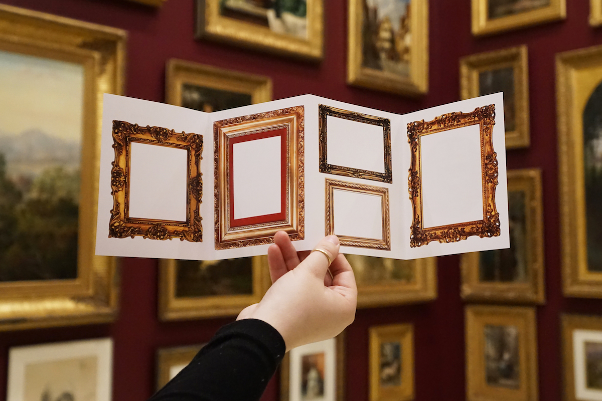 Inside a sketchbook, gold frames are empty, ready for drawings. A hand holds up the accordion book in a gallery with deep red walls filled with paintings in similar gold frames hung floor to ceiling.