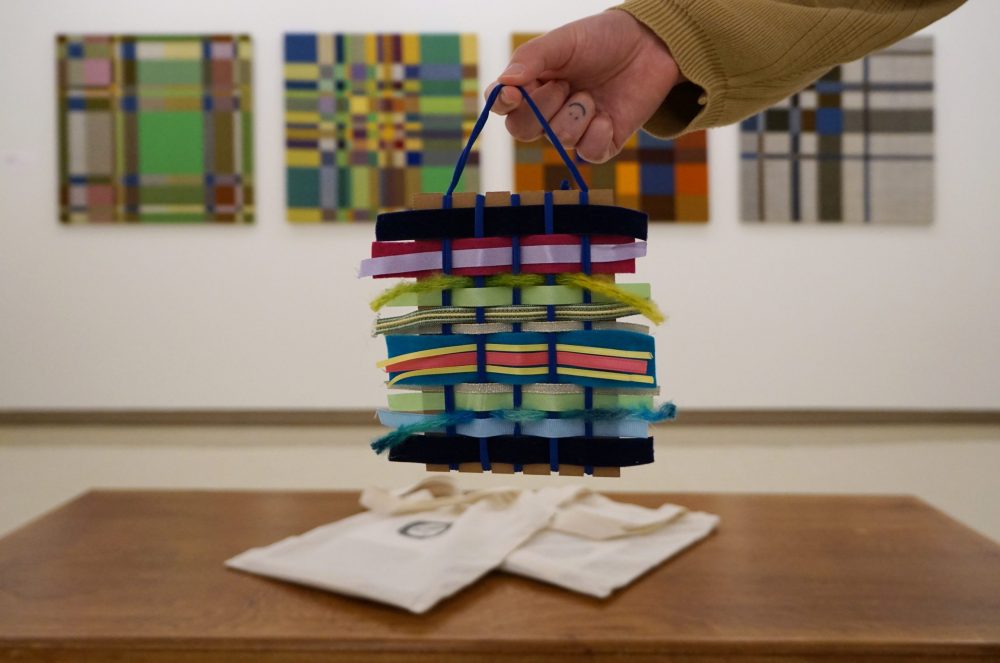 A hand holds a small weaving made of yarn and ribbon of many different colours, textures and thicknesses. Below on a bench rests two canvas bags. In the background, large weavings of colourful grid hang on a white wall.
