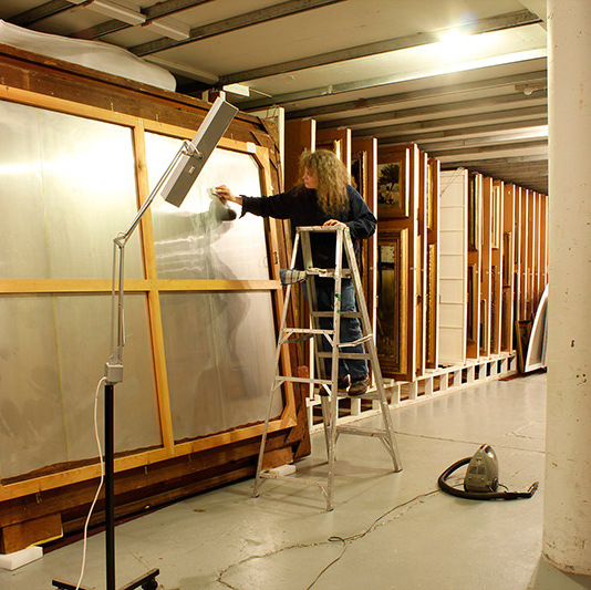 Jane Tisdale, Owens Art Gallery Fine Art Conservator stands on a ladder while cleaning the back of a very large framed painting in the painting vault.