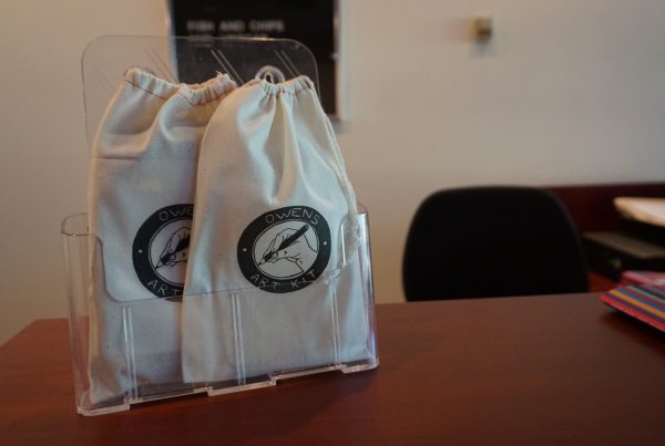 Two canvas drawstring bags sit in a clear plastic display shelf on a wooden desk. Printed on the bags is an illustration of a hand holding a pencil encircled by a black band with text that reads "OWENS ART KIT".