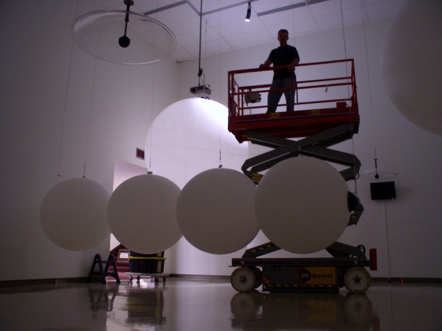 Roxie Ibbitson, Owens Art Gallery Registrar/Preparator stands silhouetted atop a scissor lift, high above a large installation of weather balloons.