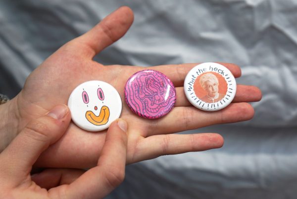 A hand holds up three pin-back buttons. One is a smiling clown face, the next a tangle of pink worms and th elast had an adult's face with the text "what the heck" written above them.