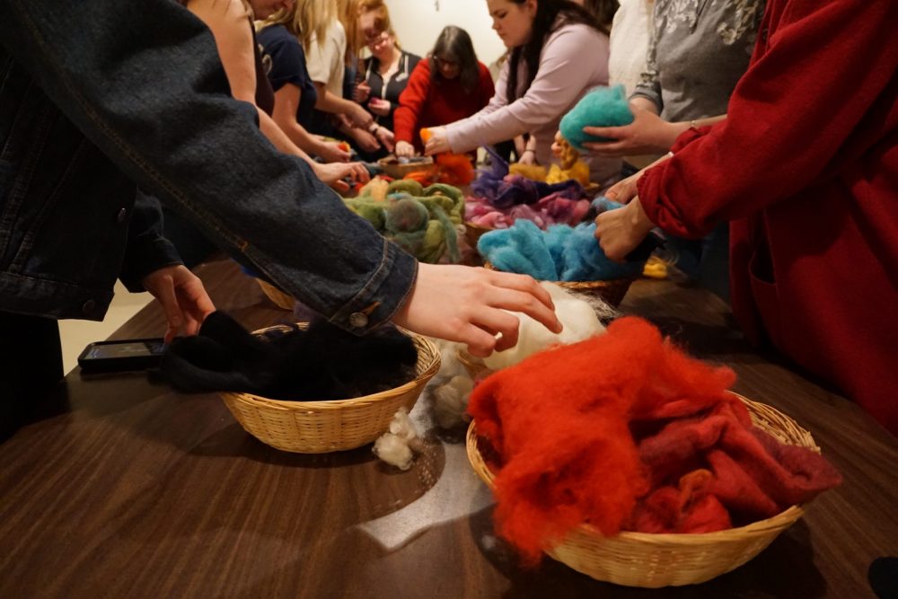 People crowd around a long table, reaching out to select colours from overflowing baskets of soft wool roving.