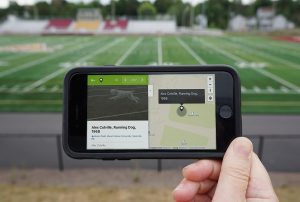 A smartphone is held up in front of a lush green sports field. On the right side of the screen is a map with a point that reads, "Alex Colville, Running Dog". On the left of the screen is an image of a Wire Fox Terrier racing across a sports field marked with painted white lines.