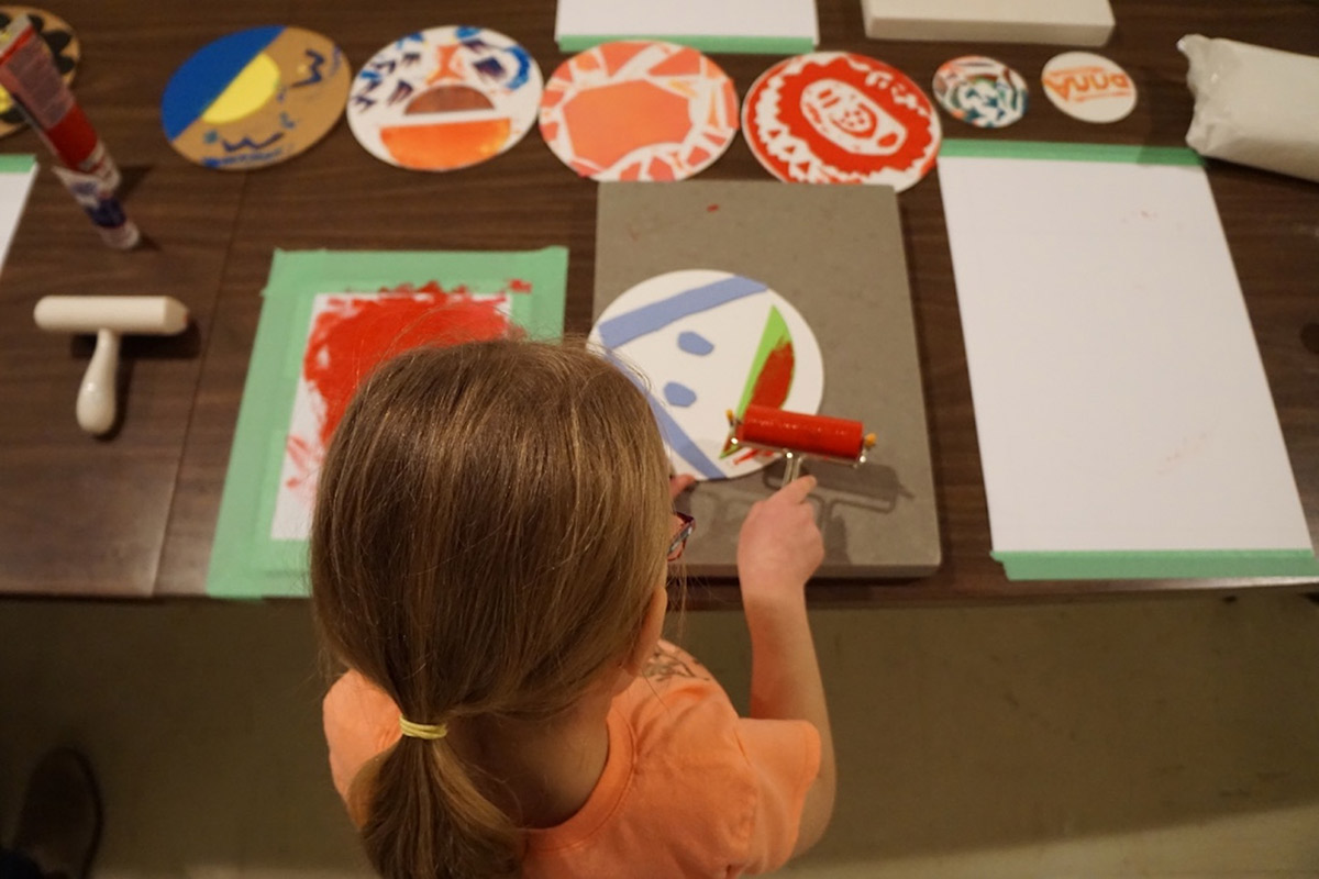 A child stands over a table inking-up a circular printing plate with face-like features. In their hand they hold a rubber roller with red ink. On the table there is a row of similar round plates that feature colourful hand-cut squiggles and shapes.