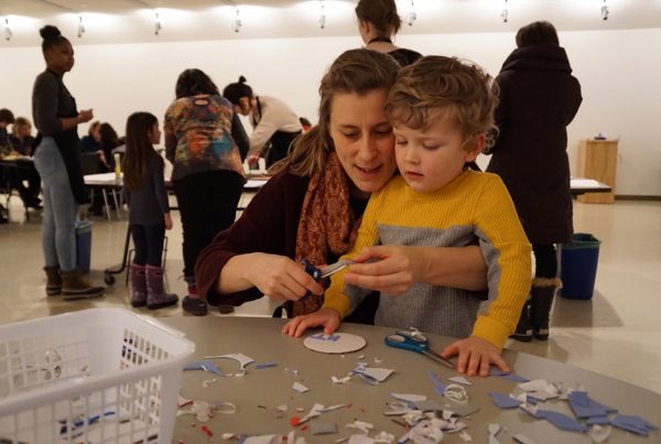 In a gallery an adult and child sit together at a table cluttered with small scraps of cut paper. The adult uses a pair of scissors to cut small pieces of paper while the child watches. Behind them adults and children work at other tables.
