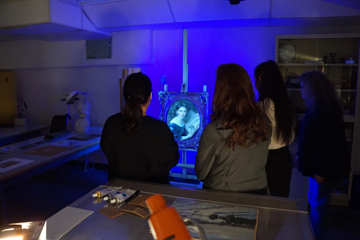 A group of adults stand in an art conservation lab examining a framed portrait painting under ultra violet light. The whole scene is cast in a blue glow.