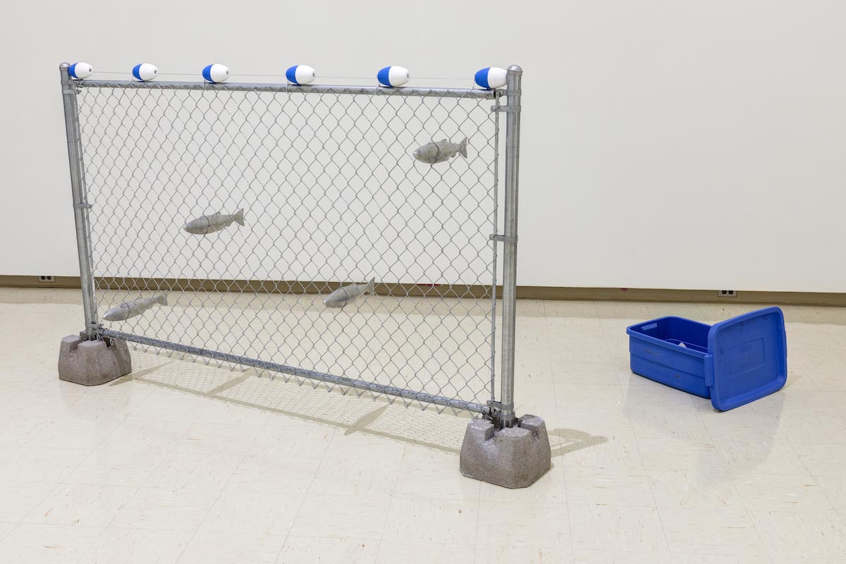 Four aluminum Arctic Char are caught in a fishing net sculpture built from a section of chain-link fence, held upright by cinder blocks. Along the top of the fence are 6 blue and white buoys. On the floor next to the fence is a blue Rubbermaid bin.