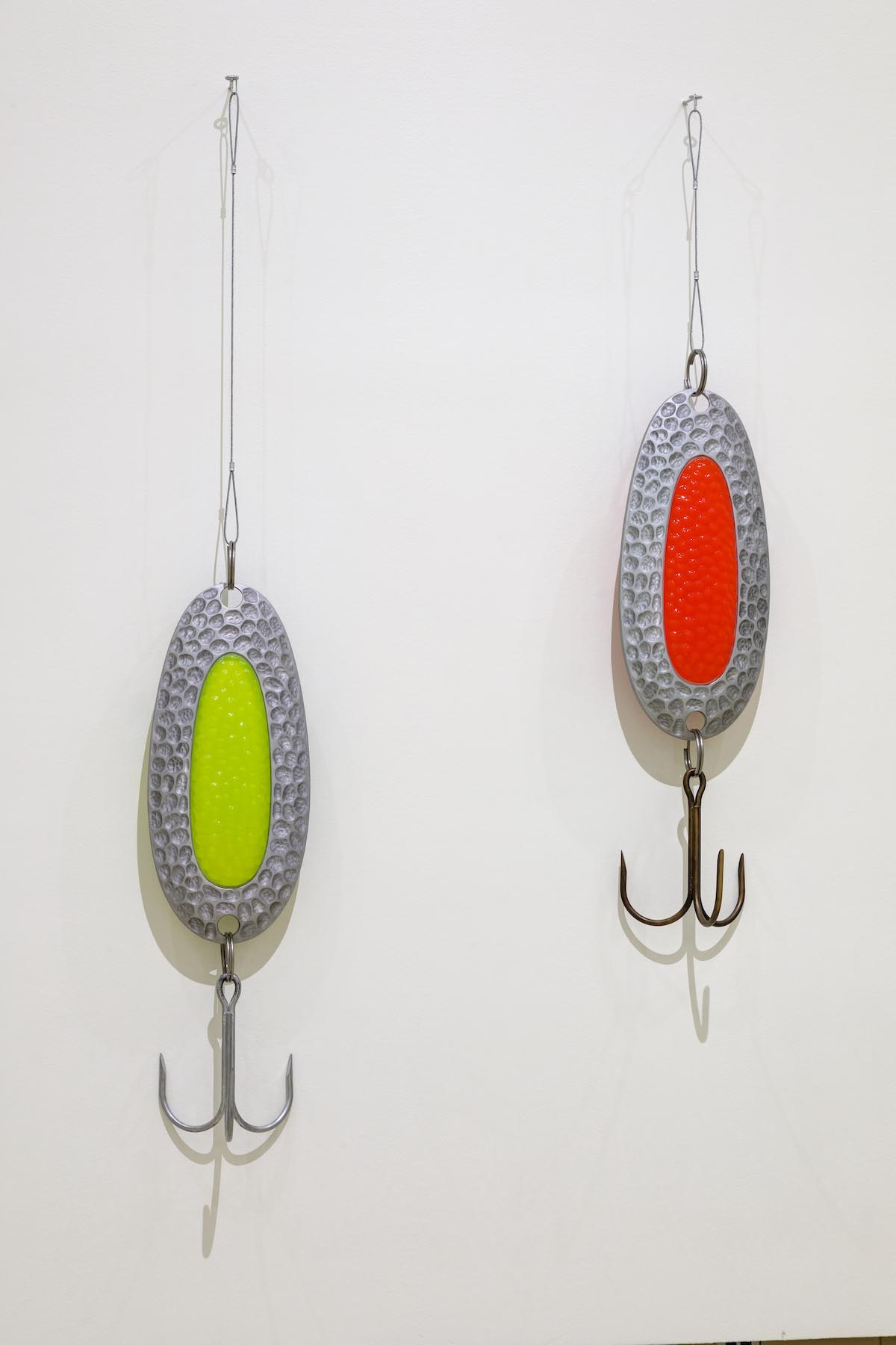 Two oversized spoon fishing lures, each measuring over five feet tall, are installed on a white gallery wall. Each has a hook attached. One has a fluorescent green centre and the other red.
