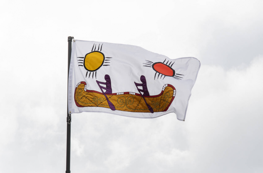 A brightly coloured flag flies against a cloudy sky. The image on the flag features two suns, one yellow and one red, and two figures paddling a canoe.