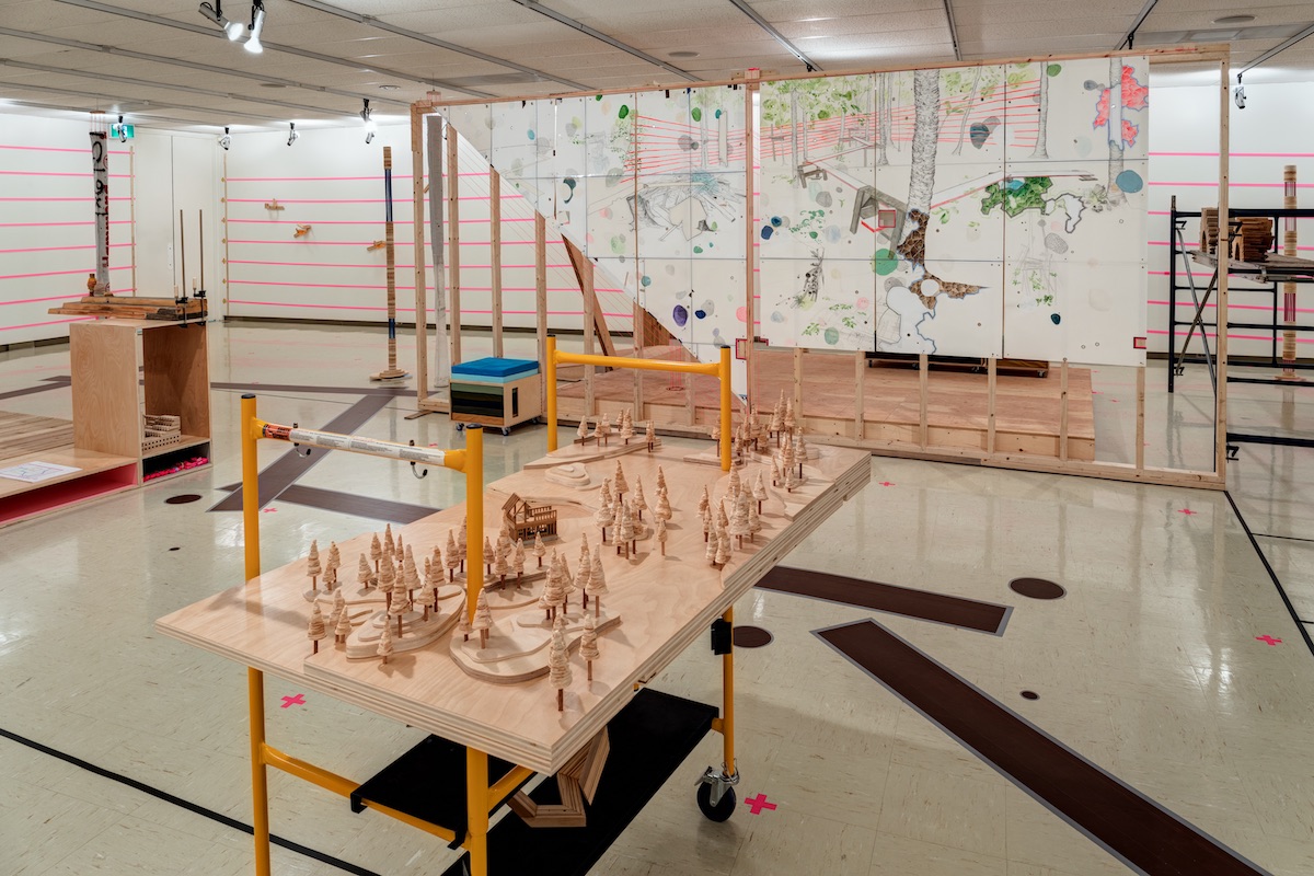 An installation fills a gallery. In the foreground a small forest of wooden trees sit on top of a sheet of pale wood on a construction table with yellow legs. Behind it, an unfinished wall of wooden beams stands from floor to ceiling. Large drawings of trees and abstract shapes rendered in graphite and bright coloured ink on paper complete the wall. Bright pink strips of tape run in parallel horizontal lines throughout the installation, connecting sculptures.