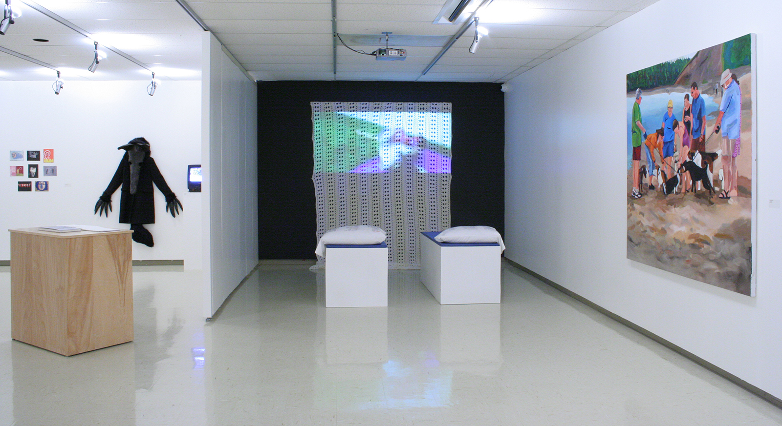 In a gallery various artworks are on display, including a black bird-like coat, a video screen, a lace curtain with a video projection, and a large painting of people with dogs on a beach.