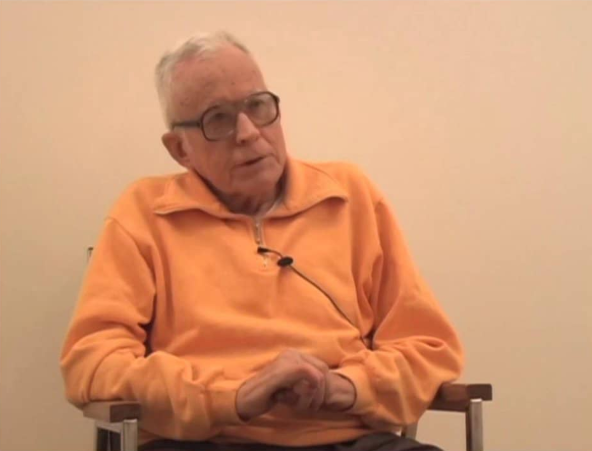 The artist, Glenn Adams sits in a chair. He is wearing an orange sweater, lapel microphone and thick-rimmed glasses.