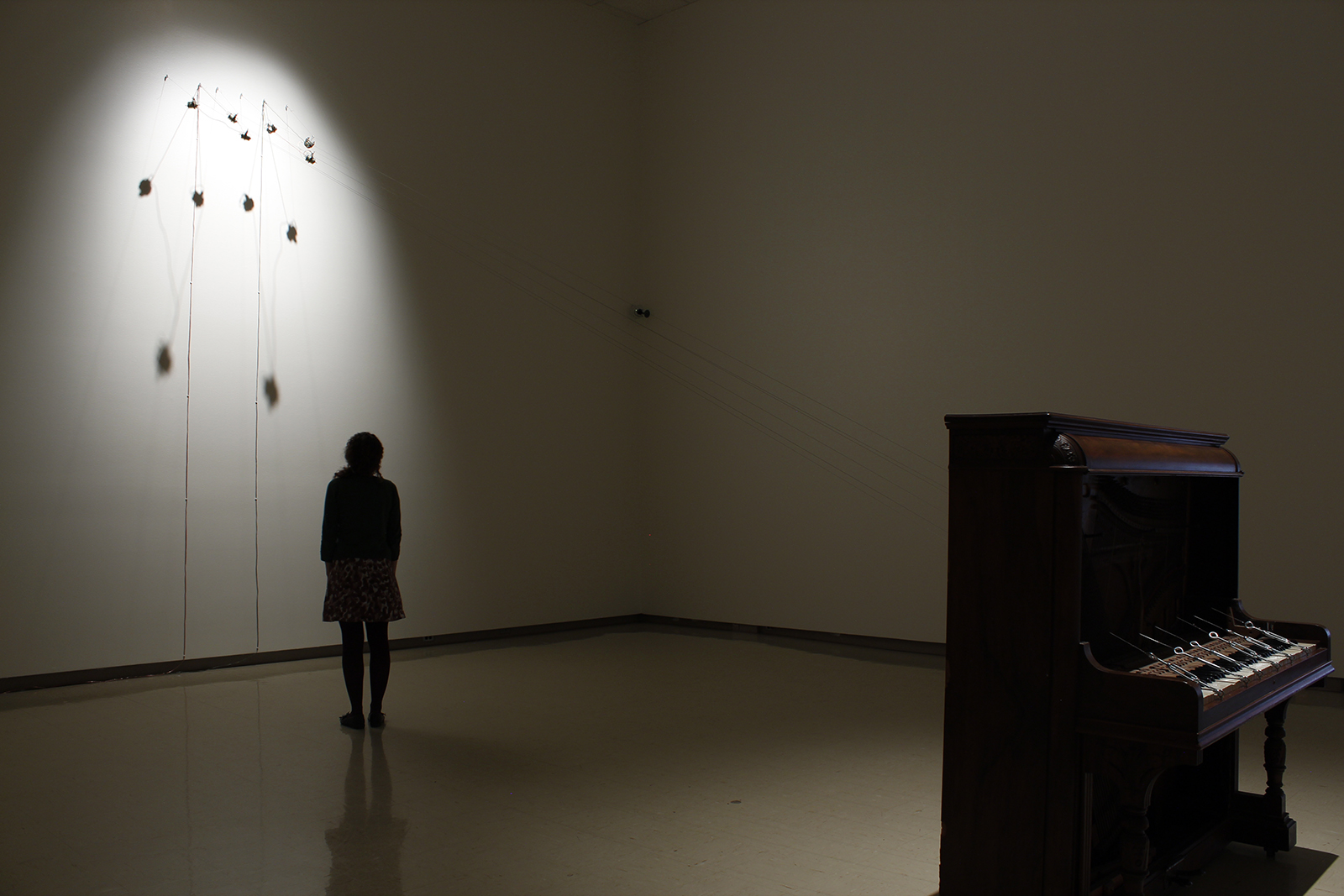 In a darkened gallery space, an adult looks up high on the wall to strings which are connected to a standing piano.