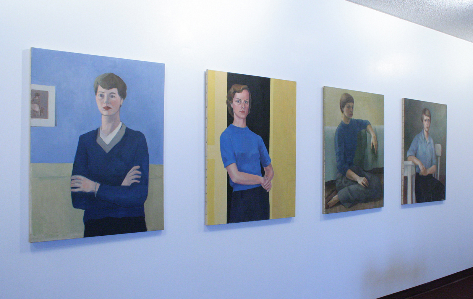 Four painted self-portraits of three women and one man hang on a white wall next to two small black and white photographs.