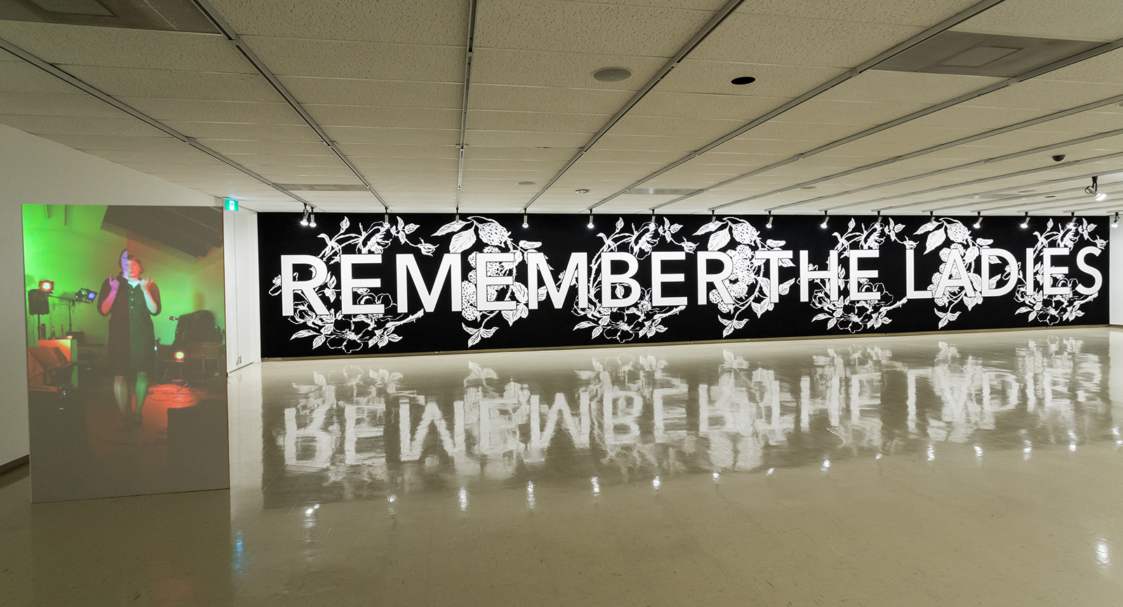 A long wall flocked with black velvet reads "Remember the Ladies" in bold font surrounded by raspberry brambles. In front of the wall, a video of a figure plays with distorted red and green colours.