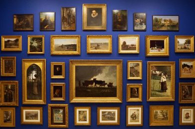 Framed paintings and works on paper are hung salon-style on a deep blue wall. Rows of gold and decorative frames in various sizes are displayed from floor to ceiling.