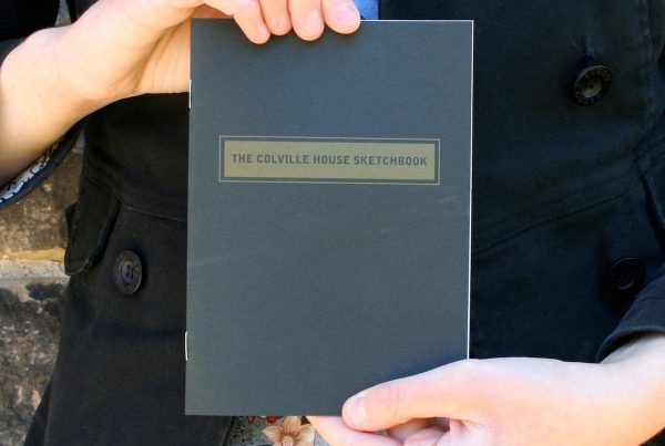 A pair of hands holds up a small, staple-bound book with the cover facing outwards. The book's cover is black, and a gold rectangle across the top contains the title “THE COLVILLE HOUSE SKETCHBOOK”.