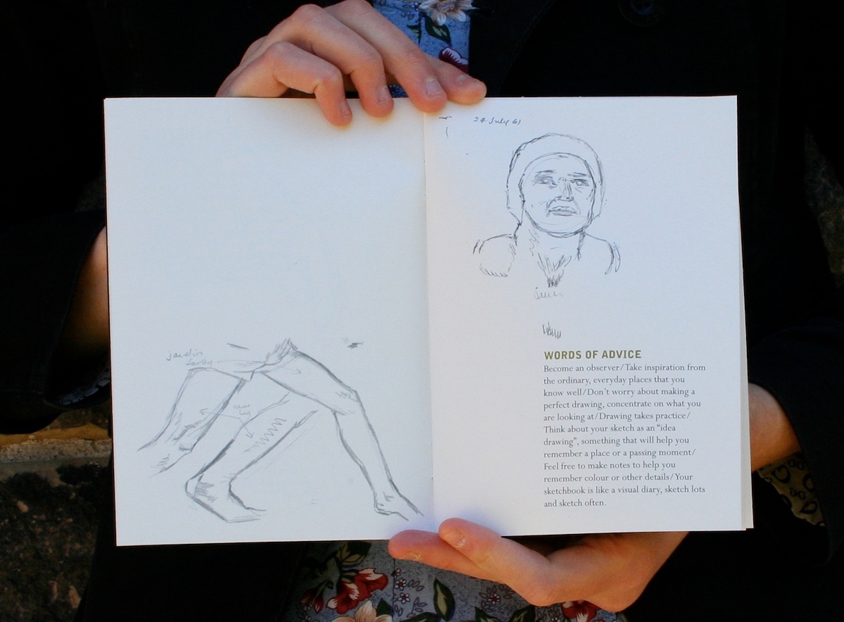 A close-up on a pair of hands holding an open book with the pages facing outwards. The pages show scans of pencil drawings of legs and a person's head. In the bottom right corner of the right page, there is a short paragraph titled “WORDS OF ADVICE”.