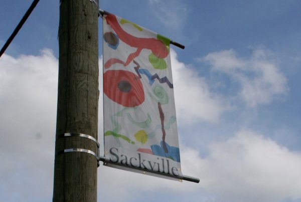 A vertical banner attached to a telephone pole is viewed from below. The banner shows an image of colourful shapes including circles, zigzags and swirls against a white background with the word "Sackville" printed across the bottom.