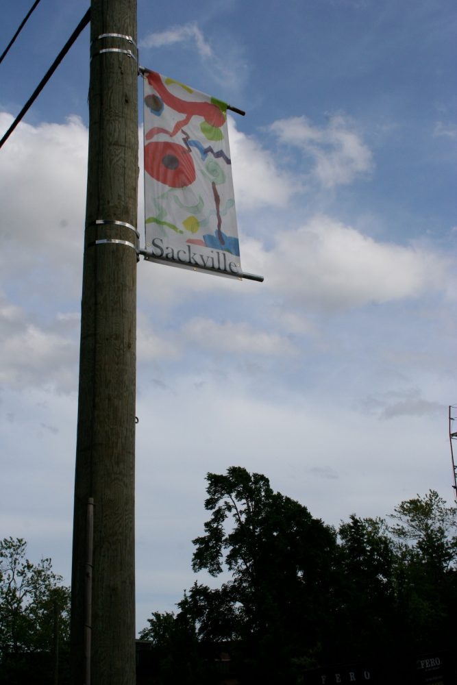 A vertical banner attached to a telephone pole is viewed from below. The banner shows an image of colourful shapes including circles, zigzags and swirls against a white background with the word "Sackville" printed across the bottom.