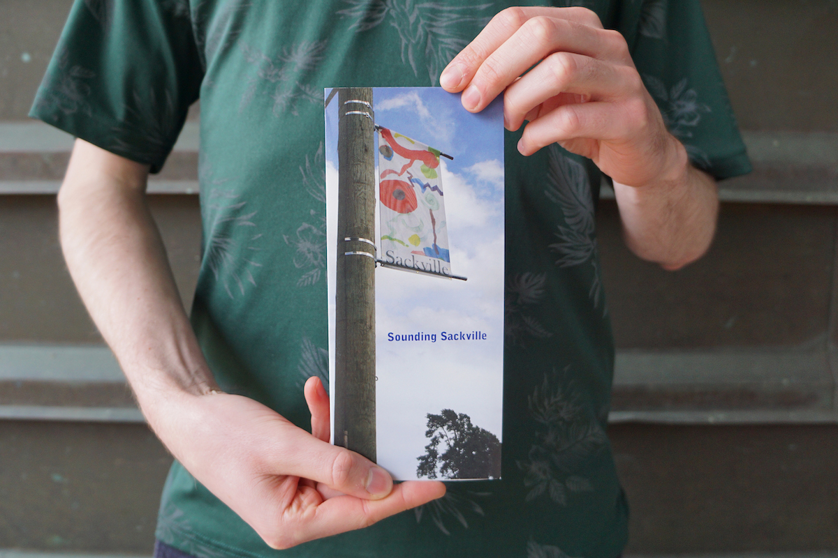 A pair of hands holds up a small publication with the title "Sounding Sackville” printed in blue letters. The cover features a photograph of a colourful banner with geometric shapes installed on a telephone pole with the word “Sackville” printed across the bottom.