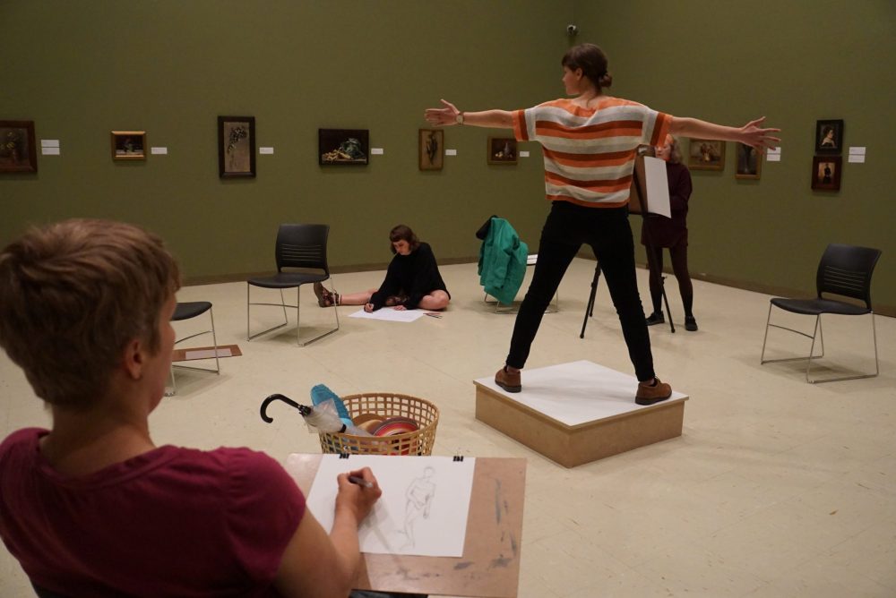 A clothed figure model stands in the centre of a gallery on top of a wooden plinth, with their feet apart and arms outstretched. Adults sit in a circle around the model, drawing them from observation.