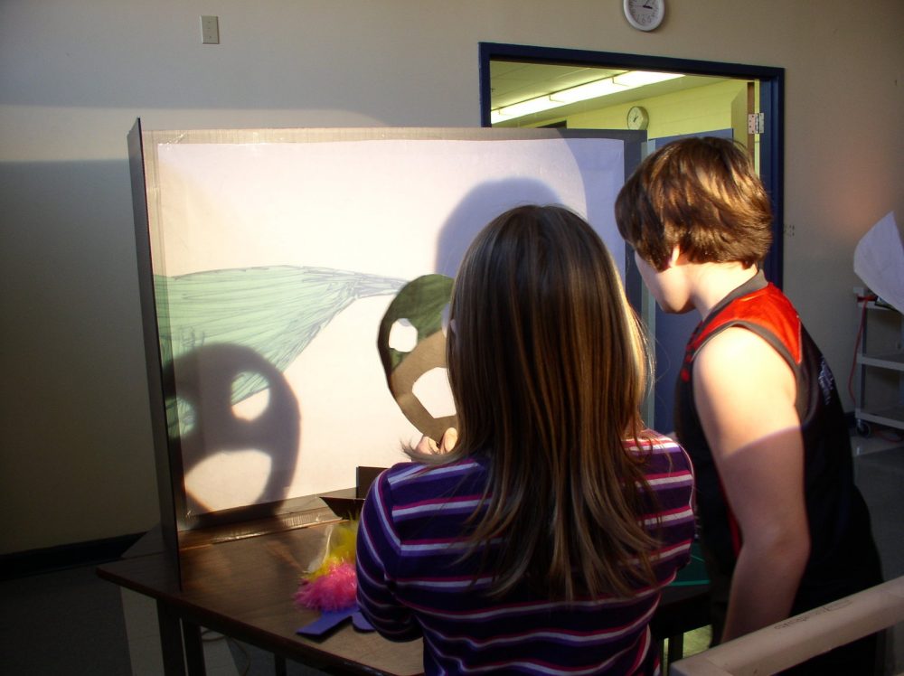 Two children stand in a darkened room in front of a small screen with a projected drawing. One of them holds a paper smiley face that casts a shadow of the smiley face onto the screen.
