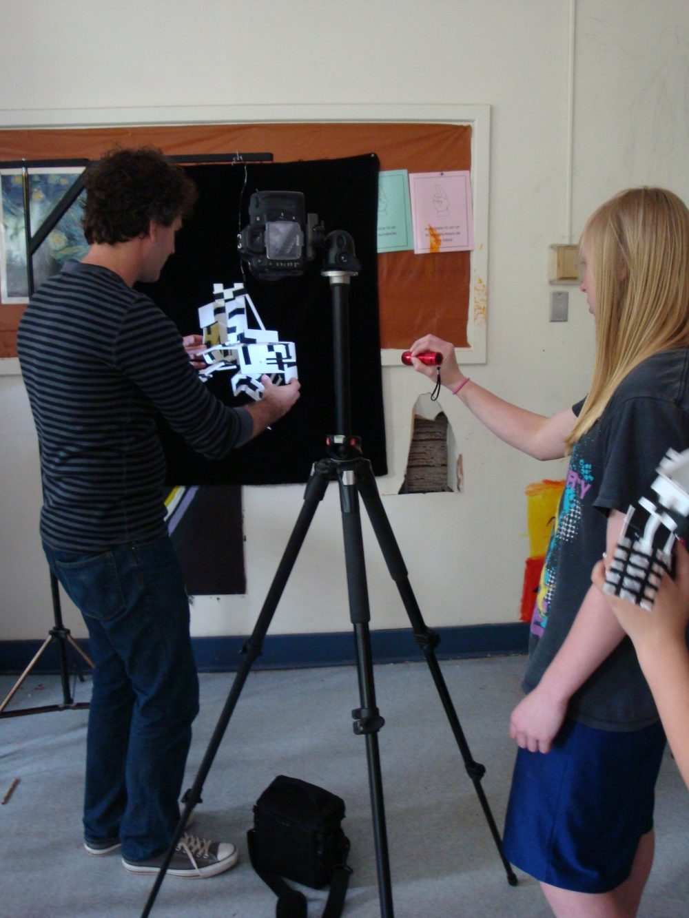 Ryan Suter holds a small satellite-like object against a black backdrop while a young person illuminates it with a flashlight. A camera on a tripod is positioned between the two people.