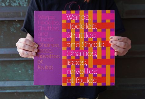 Hands remove a publication from a slip case. The cover reads "Warps, Heddles, Shuttles and Sheds" in pink and "Chaîne, Līsses, navettes et foules" in orange, on the purple cover. On the slip case the same text appears in white on top of woven bands of vibrant orange, pink and red against a purple background.