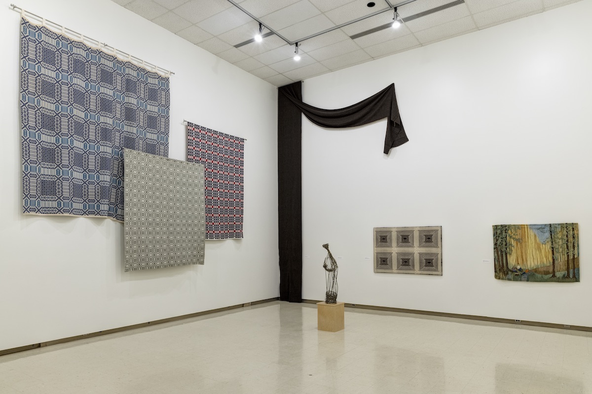 Two walls in a brightly lit gallery display woven art works. On one wall, three large square weavings hang together in a group, partially overlapping one another. On the opposite wall, a long thin weaving hangs from floor to ceiling and then drapes partway across the top of the wall. To the right, a second smaller weaving and a woven tapestry are displayed on the wall. In the middle of the gallery, a bronze statue of delicately woven reeds in the form of a human stands on a plinth.