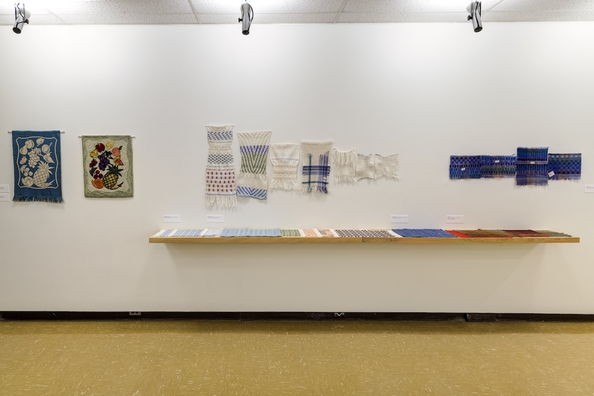 Groupings of woven artworks are hung across a white wall in a brightly lit gallery. Below these works more weavings are displayed on a long wooden shelf.