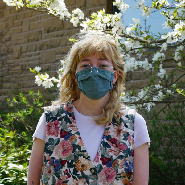 Portrait of Emma Connors standing outside on a sunny day wearing a blue cloth face mask, glasses and floral vest over a white t-shirt.