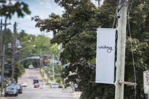 On the right side of the image is a grey banner installed on a telephone pole with cursive text that reads “waiting”. In the background is a tree-lined street dotted with parked cars.
