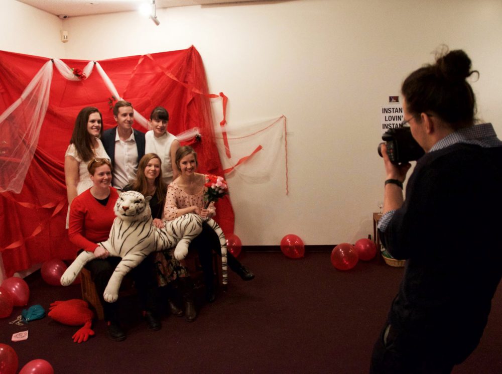 In the lobby of the Owens Art Gallery, a group of smiling adults gather together in front of a backdrop decorated with red fabric, streamers, balloons, and plastic flowers. Three adults stand while three sit in front, holding a large white stuffed tiger. A photographer stands before them, ready to capture their photo using a Polaroid camera.