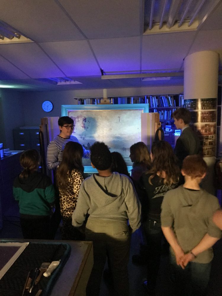 In a darkened room, an adult stands in front of a framed artwork lit by blue ultra violet light and addresses a group of children. The adult extends their arm towards the work, pointing something out to the group.
