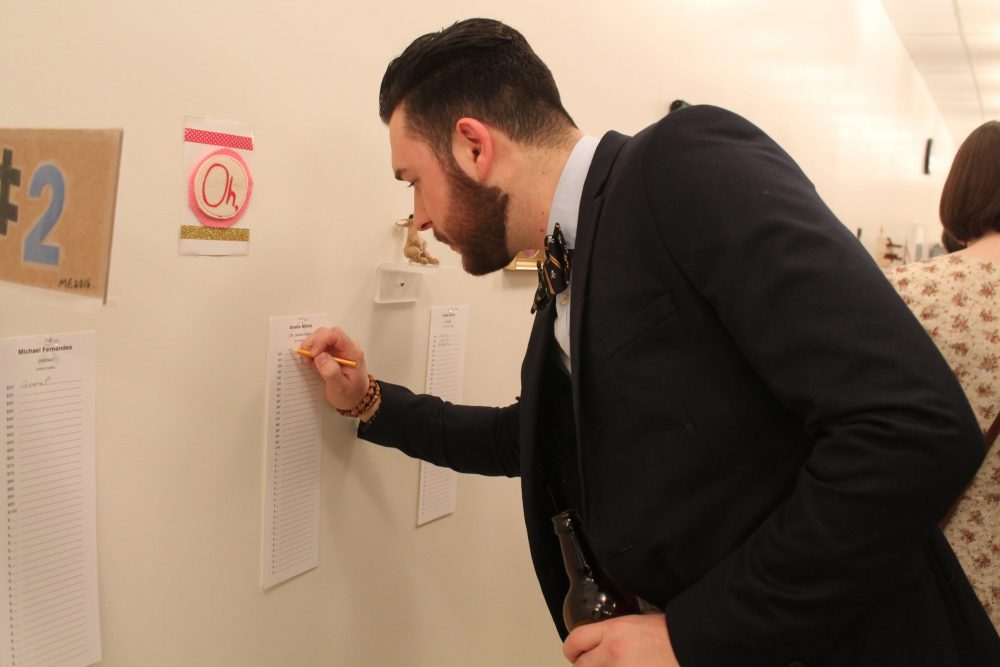 An adult leans to write on a sheet of paper beneath an artwork. The pink and white felt patch with the word “Oh,” printed on it, is one of many small artworks displayed in a horizontal row on a wall.
