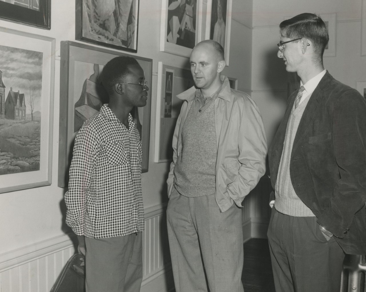 A black and white photograph of three men standing in a gallery. Two rows of paintings hang on a wall behind them.