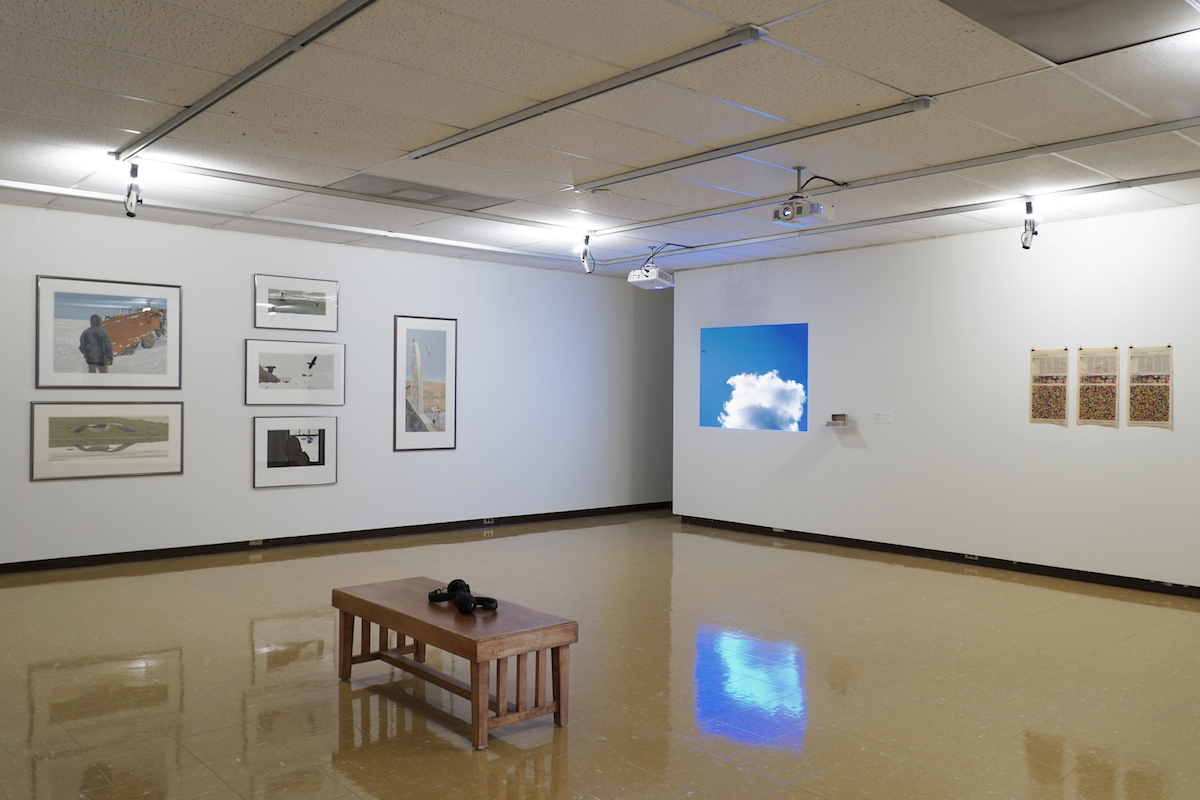 In a gallery, on the left wall six silkscreen artworks hang on a white wall, each in a silver frame. To the right, an image of a cloud is projected onto the wall next to three pages of newspaper crosswords.
