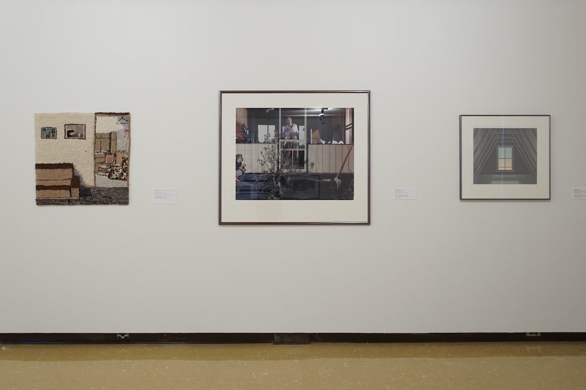 Three artworks hang on a white gallery wall. On the left, is a hooked rug depicting a living room. In the centre is a photograph of an adult looking out a screened porch while a child crouches to hide on the steps. The silkscreen on the right shows a window in a steeply pitched attic.