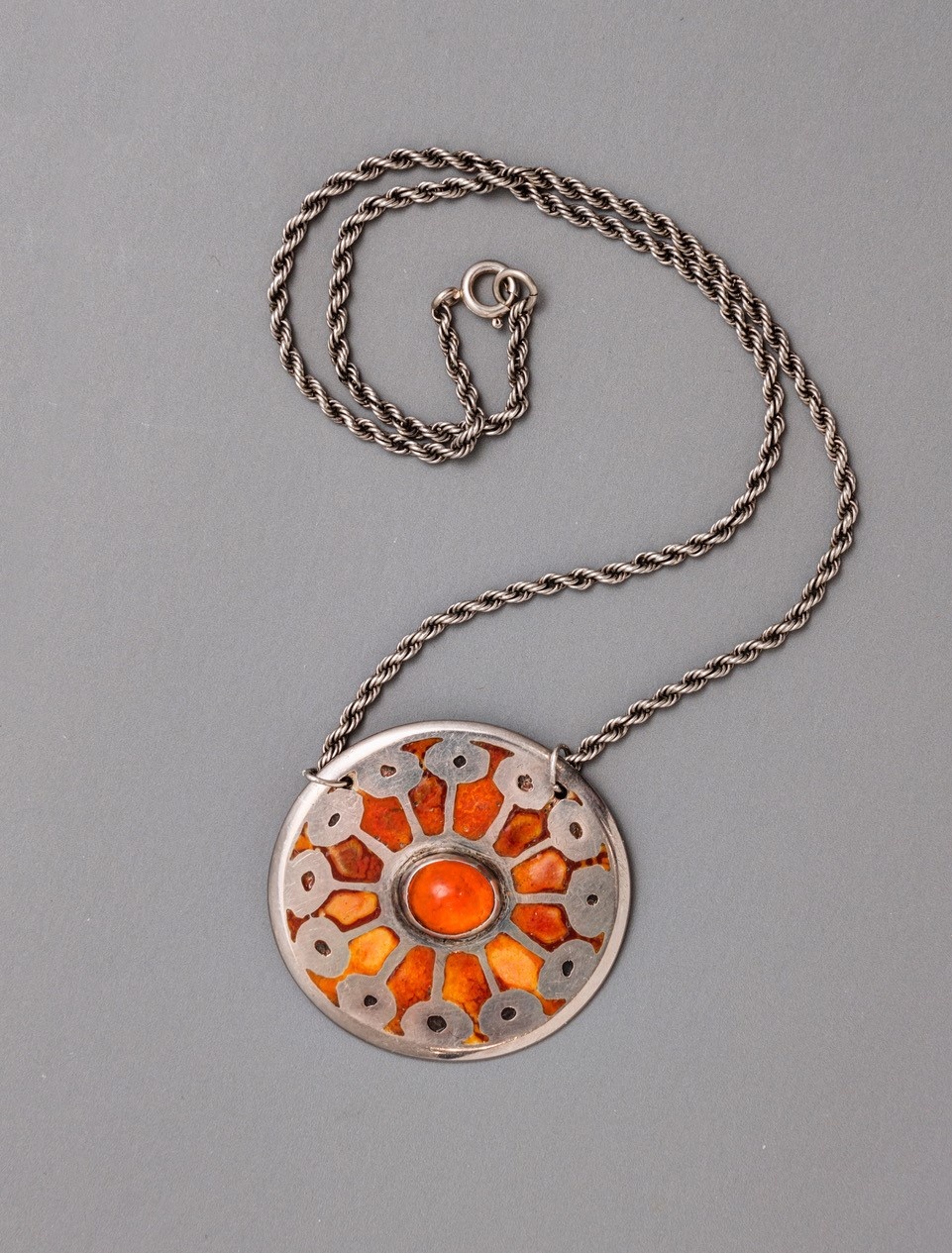 A circular silver medallion is attached to a silver chain. Silver ovals circle the outer edge of the medallion and connect to a small orange oval stone is set in the middle of the medallion with thin lines. Below the silver design is a smooth orange stone with hints of yellow and red.