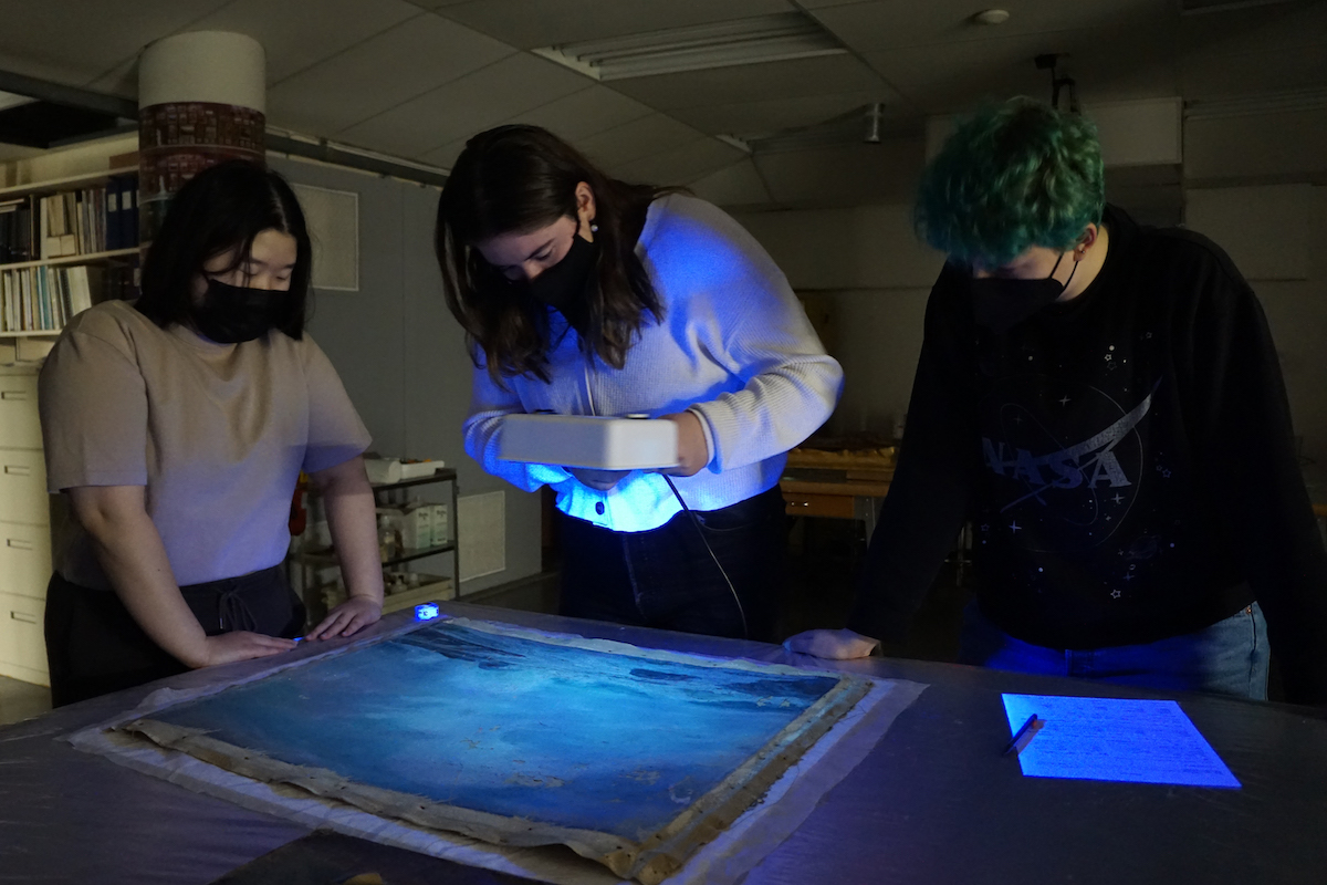 Three students stand around a painting on a table in near darkness. The student in the centre holds an Ultra Violet light, illuminating the painting with a blue glow.