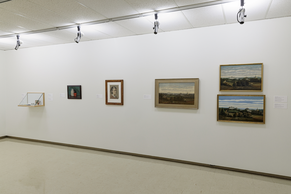 On a gallery wall, three paintings of a landscape hang next to a portrait, a painting of a vase and a display case.