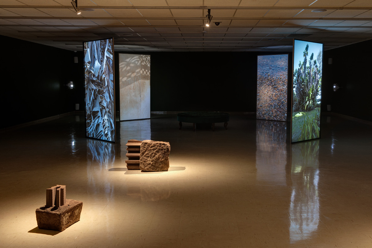 In a darkened gallery, four 8x4 foot screens are illuminated with projections of bright green foliage. In the foreground two red stone sculptures are illuminated by spot light.