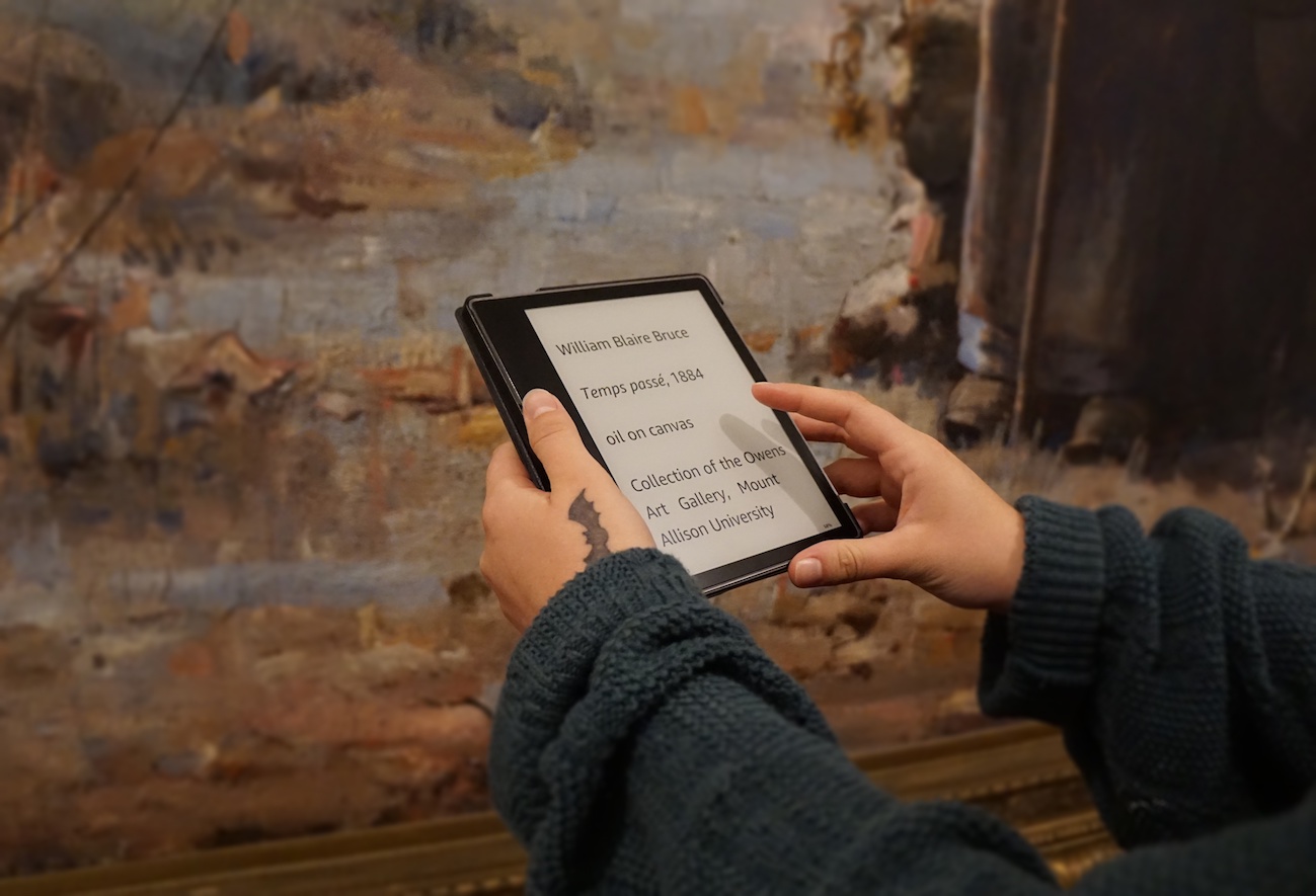 In front of a large painting, two hands hold a kindle e-reader. On the screen text reads "William Blaire Brice, Temps Passé, 1884, oil on canvas"