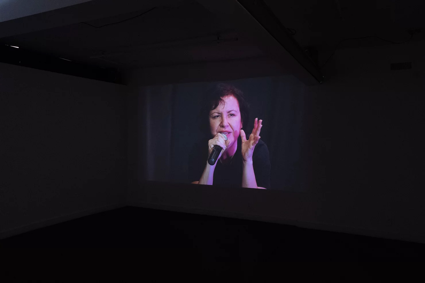 A video of an adult speaking into a microphone while gesturing expressively with their hand is projected in a darkened gallery space.