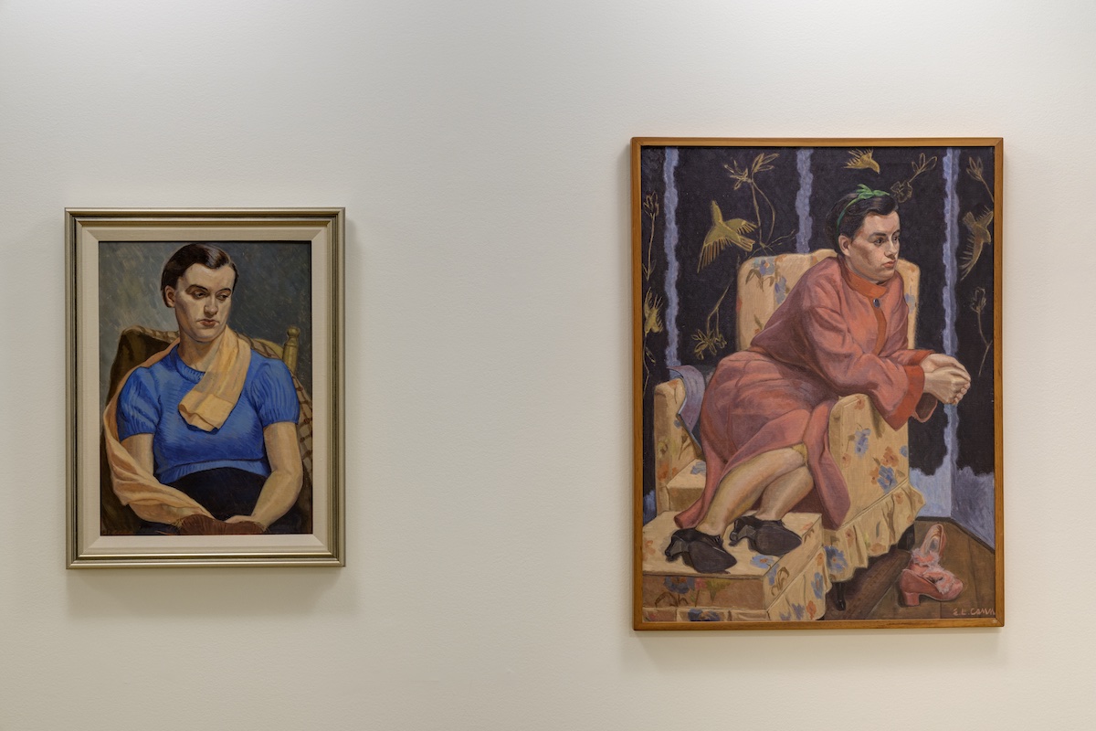 Two painted portraits hang side by side. Each depicts a woman seated in an arm chair, with solemn expressions. The woman on the left wears blue. The woman on the right wears a pink house coat and shoes as her feet rest on an ottoman. She is surrounded by elaborate purple wallpaper patterned with yellow birds.
