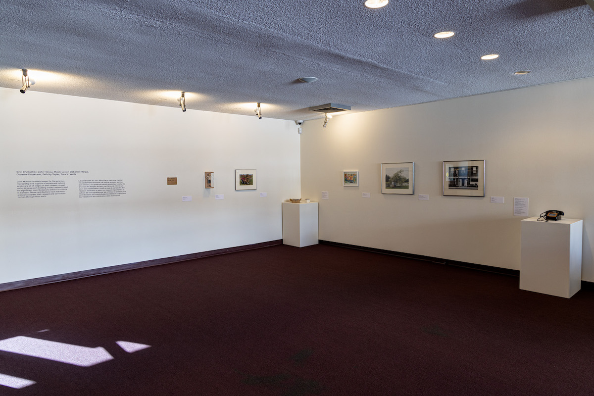 Looking into a gallery with burgundy carpet, several small artworks are displayed on two walls and plinths.